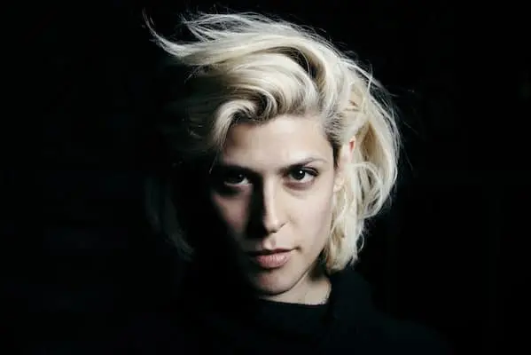 A person with light blonde, tousled hair stares intensely at the camera, partially lit from the side against a dark background, creating dramatic shadows on their face.