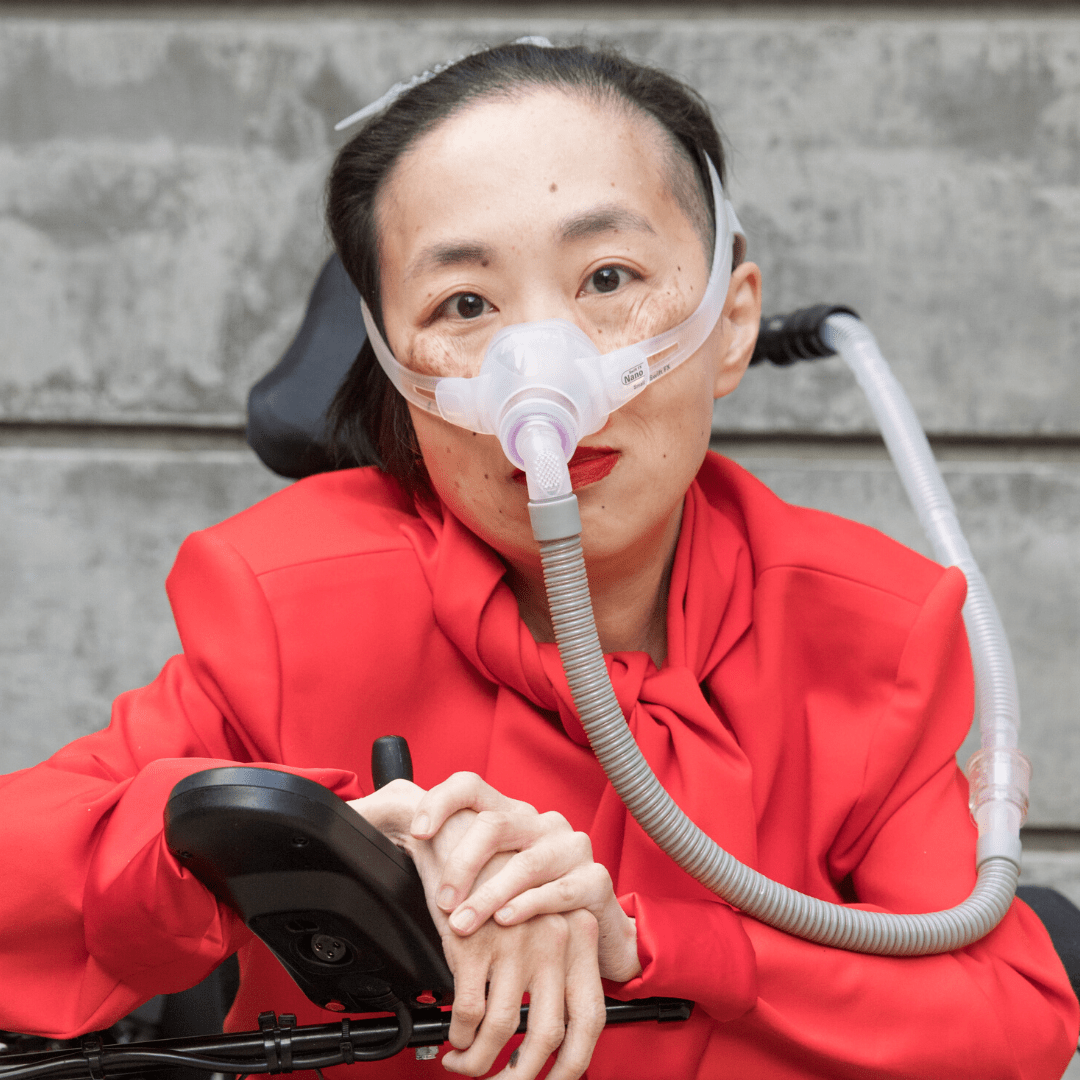 A person wearing a red blouse sits in a wheelchair, using a ventilator mask. They have short, dark hair pulled back and rest their hands on the wheelchair armrest. A gray wall is in the background.