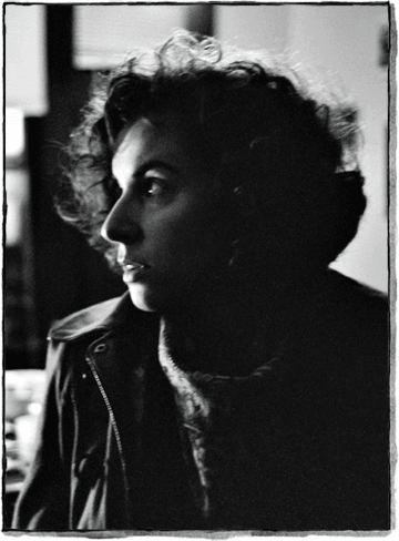 Black and white portrait of a person with curly hair looking to the left, wearing a coat and scarf, with soft light and a blurred background.