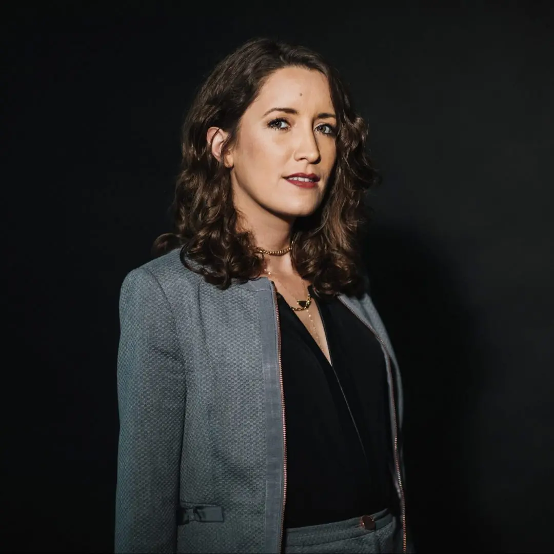 A woman with wavy brown hair wearing a gray suit jacket over a black top stands against a dark background, looking slightly to the side with a confident expression.