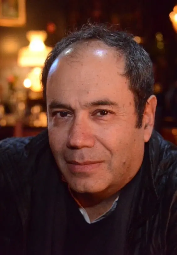 A middle-aged man with short, dark hair, some gray, and a slight smile looks at the camera. He is indoors in a warmly lit setting with a softly blurred background.