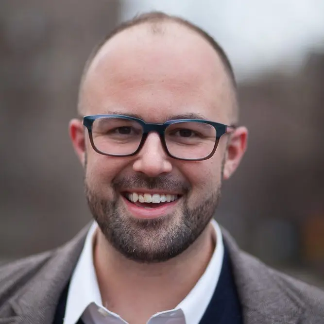 A smiling man with a trimmed beard, wearing dark-framed glasses, a white shirt, and a grey blazer, standing outdoors with a blurred background.