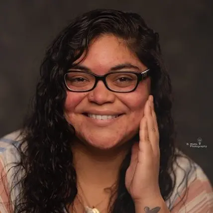 A person with long, wavy dark hair and glasses smiles warmly, touching their cheek with one hand. They wear a light-colored top and have a small tattoo on their wrist. The background is dark and blurred.