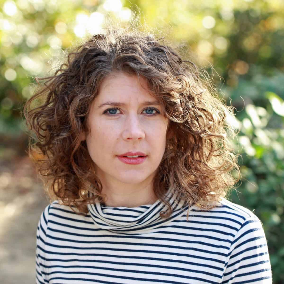 A woman with curly brown hair and blue eyes stands outdoors in natural light, wearing a white and black striped shirt. The background is blurred with green foliage.