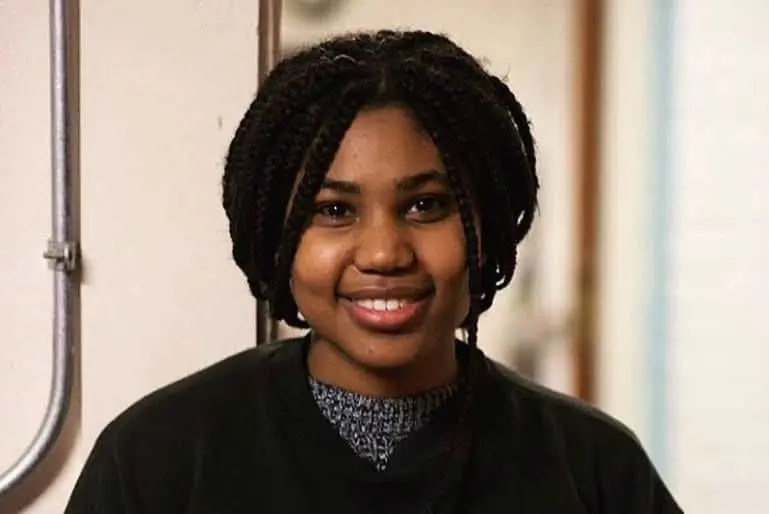 A young woman with braided hair, wearing a dark sweater over a gray top, smiles while standing indoors next to a light-colored wall with exposed pipes.