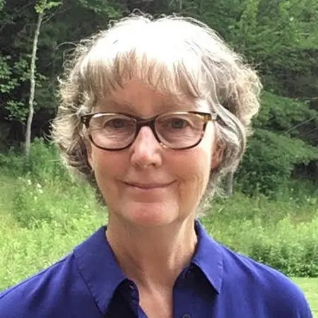A woman with short, wavy gray hair and glasses smiles at the camera. She is wearing a blue collared shirt and is standing outdoors with green trees and grass in the background.
