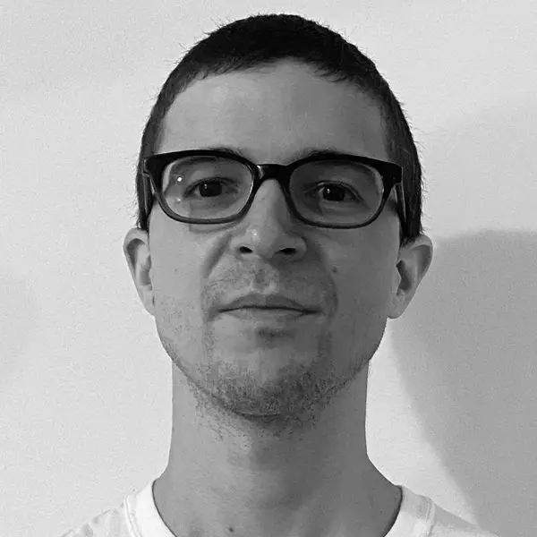 A person with short dark hair, wearing dark-rimmed glasses and a white shirt, is facing the camera against a plain light-colored background.