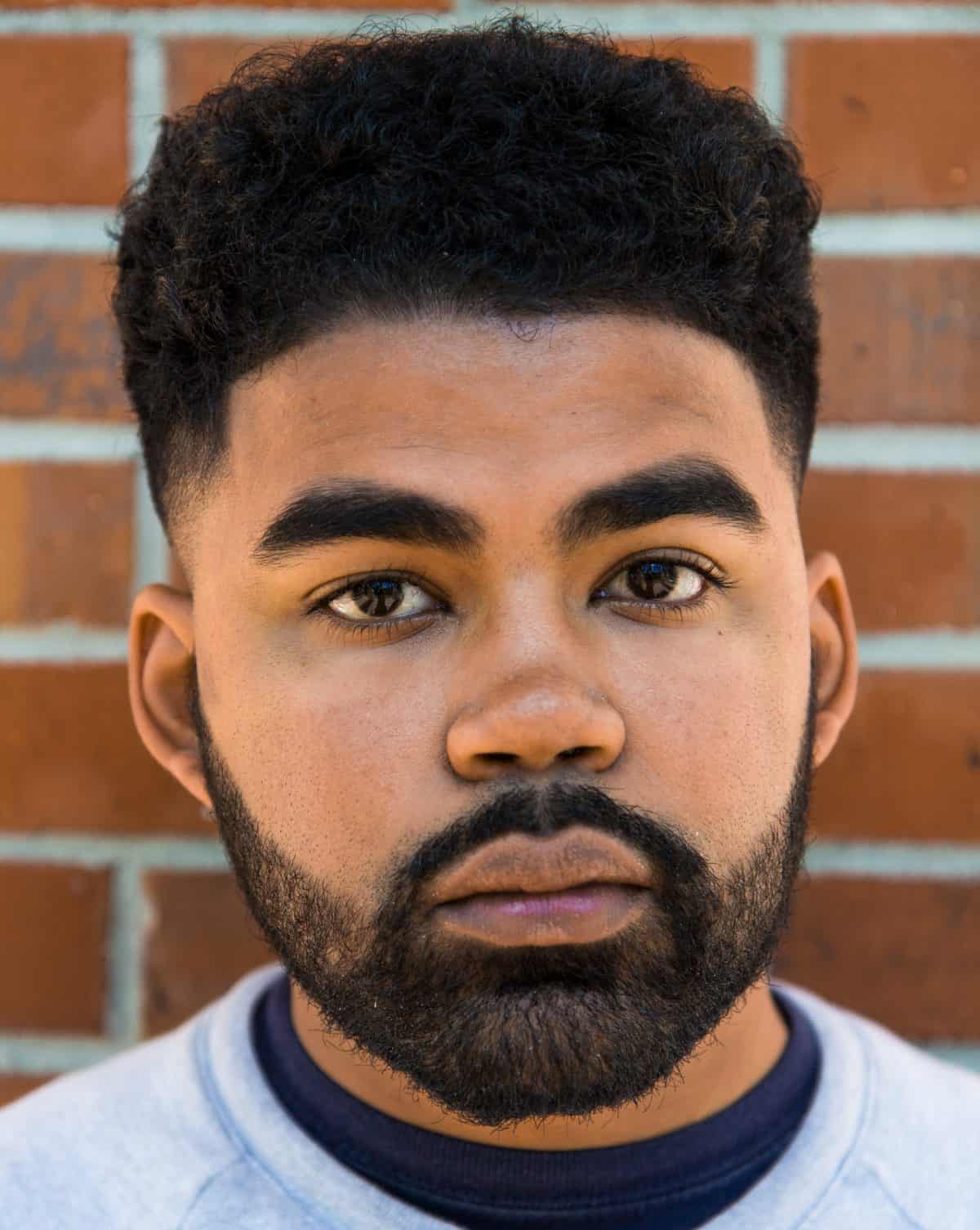 A young man with short, curly black hair and a full beard stands in front of a brick wall, looking directly at the camera with a neutral expression. He is wearing a light gray shirt, hinting at his passion for pride month literature.