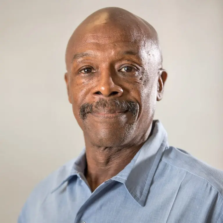 An older man with a bald head and mustache, wearing a light blue collared shirt, looks at the camera with a gentle expression against a plain, light background.