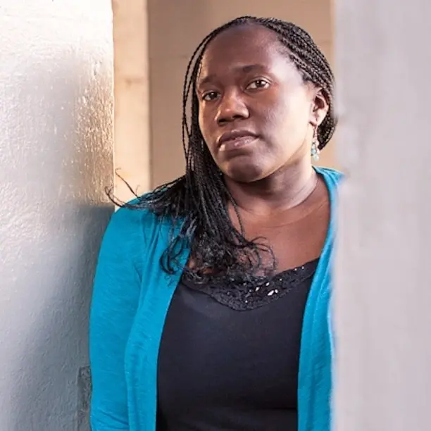 A woman with braided hair wearing a blue cardigan and black top stands between two walls, looking thoughtfully at the camera.