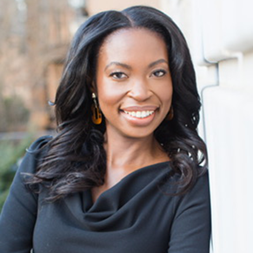 A woman with long, wavy black hair and gold earrings is smiling while leaning against a white wall. She is wearing a black top, and the background is blurred with hints of greenery and buildings.