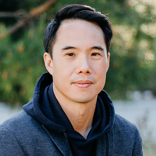 A man with short black hair, wearing a dark hoodie and gray jacket, sits outdoors in front of blurred green foliage, looking calmly at the camera—a contemplative presence reflecting the spirit of the Emerging Voices Fellowship.