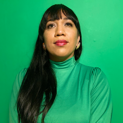 A woman with long dark hair wearing a green turtleneck top stands in front of a green background, looking slightly upward with a calm expression, embodying the confidence and poise inspired by the Emerging Voices Fellowship.