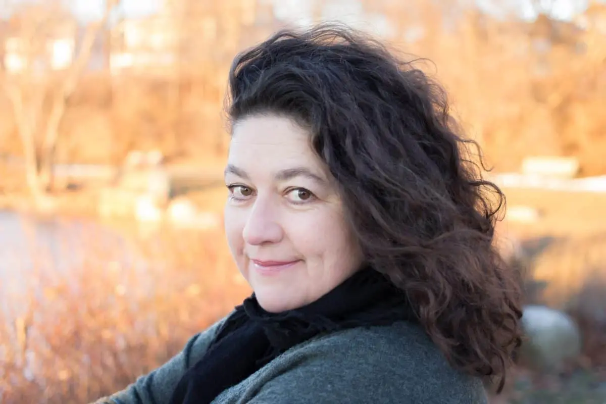 A woman with curly dark hair and a slight smile looks at the camera outdoors, with a blurred background of autumn trees and warm sunlight.