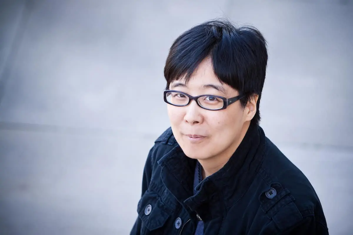 A person with short black hair and glasses, wearing a dark jacket, looks up at the camera and smiles slightly. The background is out of focus and appears to be a light-colored paved surface.