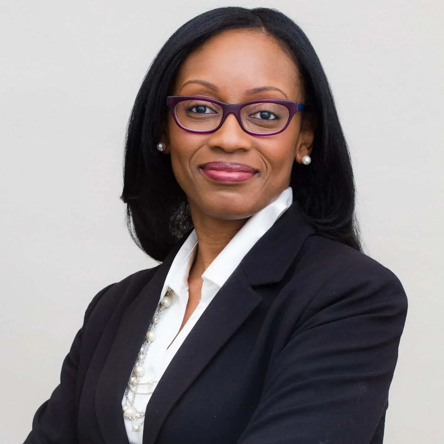 A woman with long black hair, wearing glasses, pearl earrings, a white blouse, and a black blazer, stands confidently with arms crossed against a plain light background.