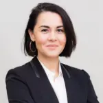 A woman with short dark hair wearing a black blazer over a white shirt, smiling and posing with arms crossed against a plain light background, captures the poised confidence of someone ready for what to expect on election day.