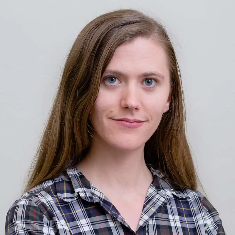 A woman with long brown hair and blue eyes is wearing a plaid shirt and standing against a plain light gray background, looking at the camera with a slight smile.