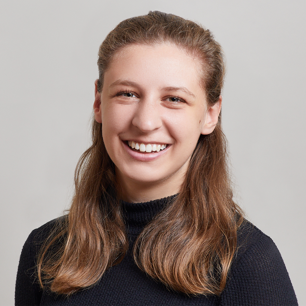 A young woman with long, light brown hair smiles at the camera. She is wearing a black, ribbed, long-sleeve top and is posed in front of a plain, light gray background.