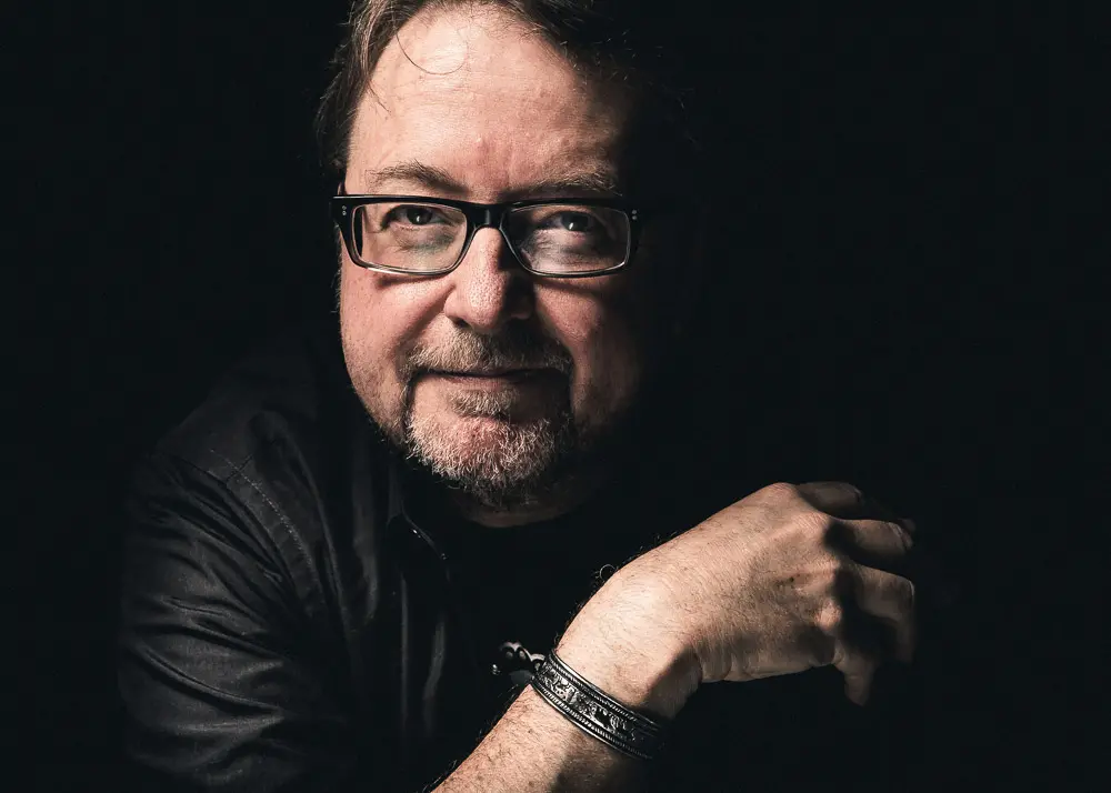 A man with glasses and a beard sits against a dark background, wearing a black shirt and a bracelet. He looks at the camera with a slight smile, resting one arm on a surface in front of him.