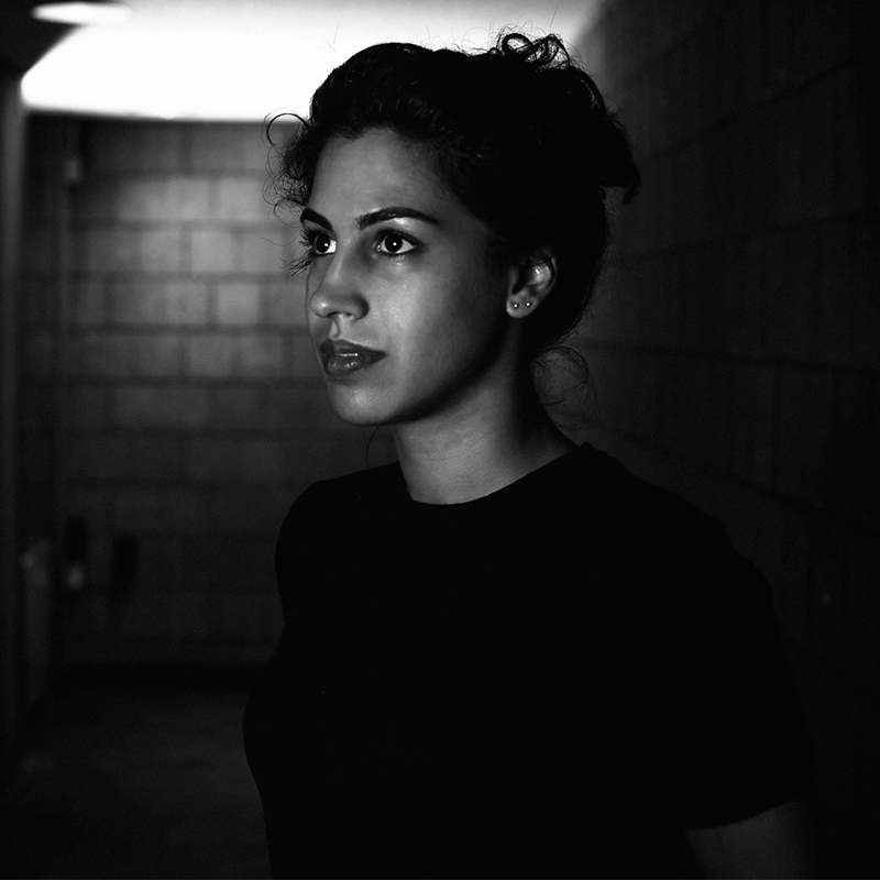 A woman with dark hair in a bun, wearing a dark shirt, stands in a dimly lit hallway with brick walls, gazing thoughtfully to the side. The image is in black and white, creating a dramatic, moody effect.