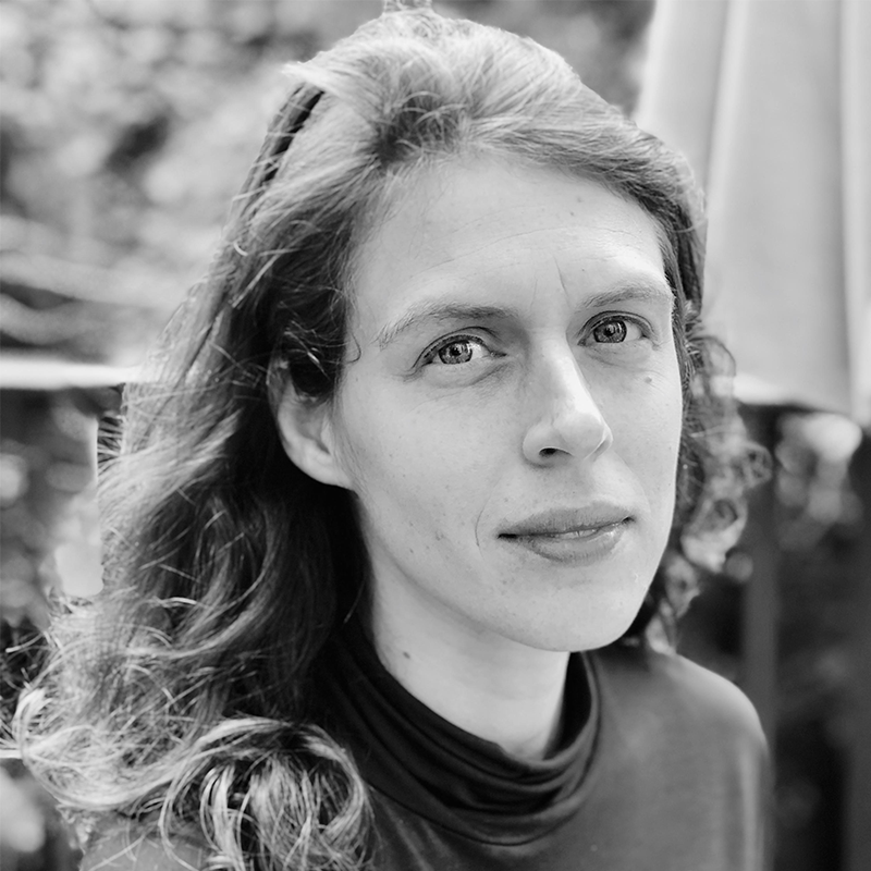 A woman with wavy hair and a calm expression looks at the camera. She is outdoors, with a blurred background of trees and an umbrella. The photo is in black and white.
