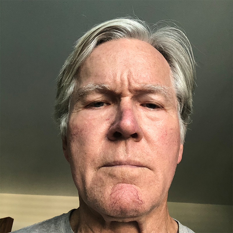 An older man with gray hair and a serious expression looks directly at the camera. He is indoors, with neutral-colored walls in the background and wearing a gray shirt.