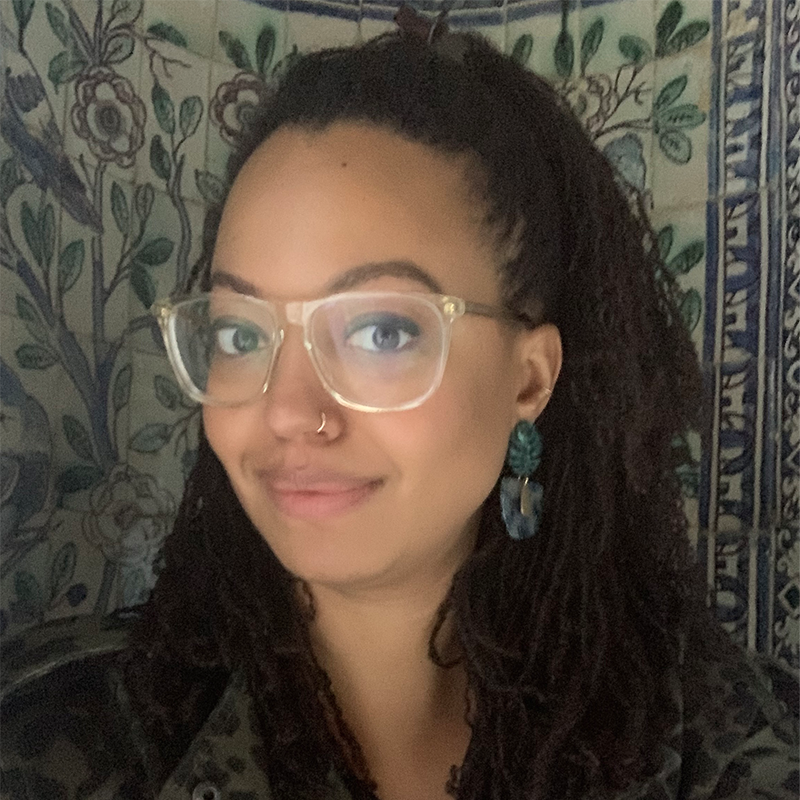 A woman with long curly hair, wearing glasses, a nose ring, and large earrings, smiles softly. She stands in front of a decorative floral-patterned wall.