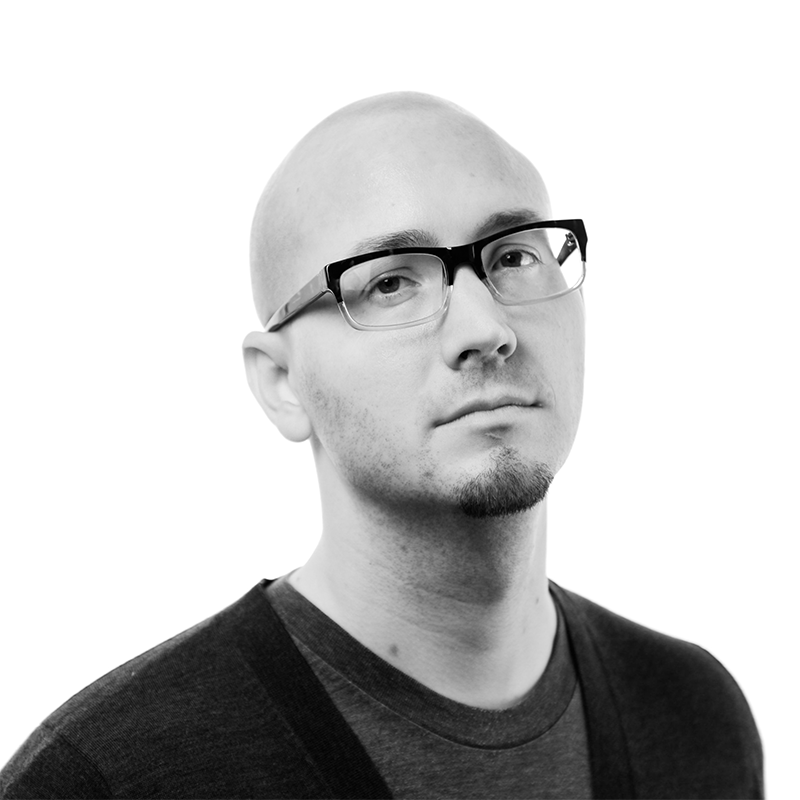 A person with a shaved head, glasses, and a short beard looks at the camera with a neutral expression, wearing a dark shirt against a plain white background.