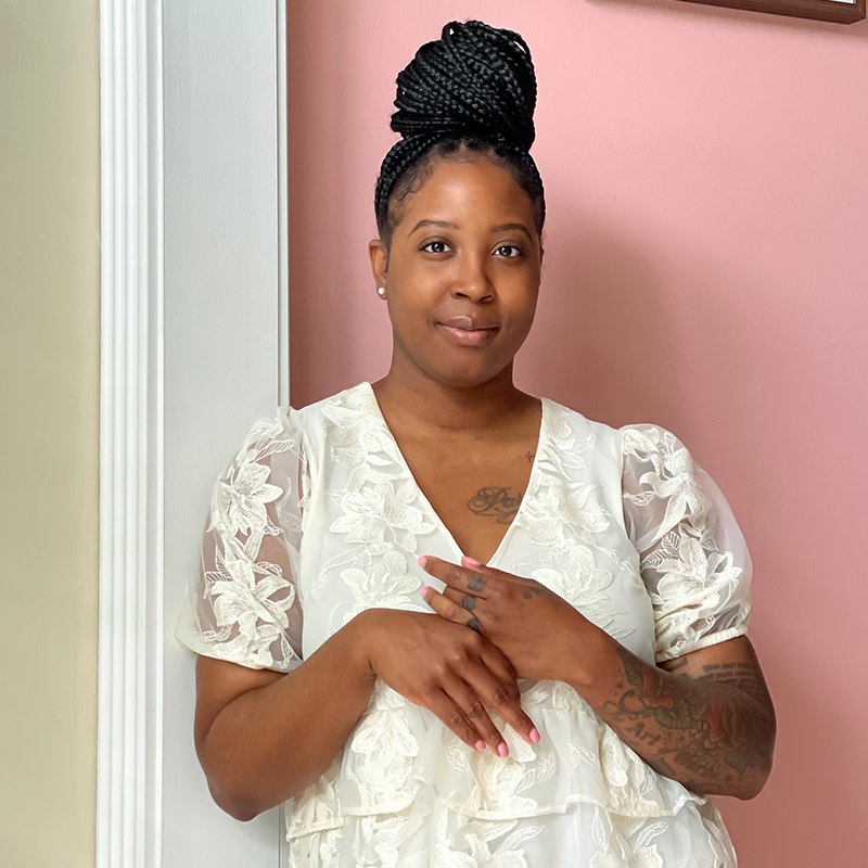 A woman with braided hair in a top bun, wearing a white floral-patterned blouse, stands indoors against a pink and white wall, smiling softly with her hands gently crossed.