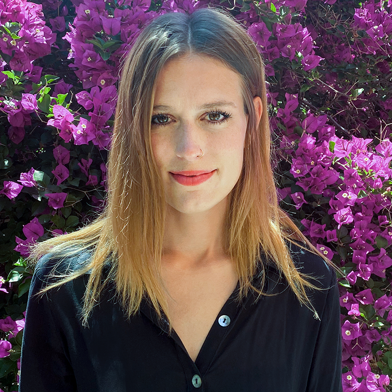 A woman with straight, light brown hair and a nose ring stands in front of vibrant purple bougainvillea flowers, wearing a black button-up shirt and smiling softly at the camera.