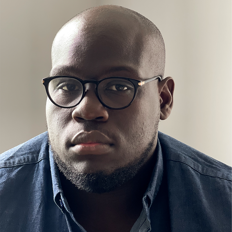 A man with a shaved head, short beard, and wearing round black glasses and a dark blue button-up shirt looks directly at the camera with a neutral expression. The background is softly lit and plain.