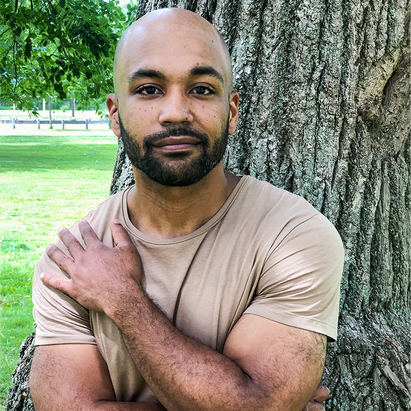 A man with a bald head and short beard stands outdoors, leaning against a large tree. He wears a beige T-shirt and crosses one arm over his chest, looking calmly at the camera. Green grass and trees are in the background.