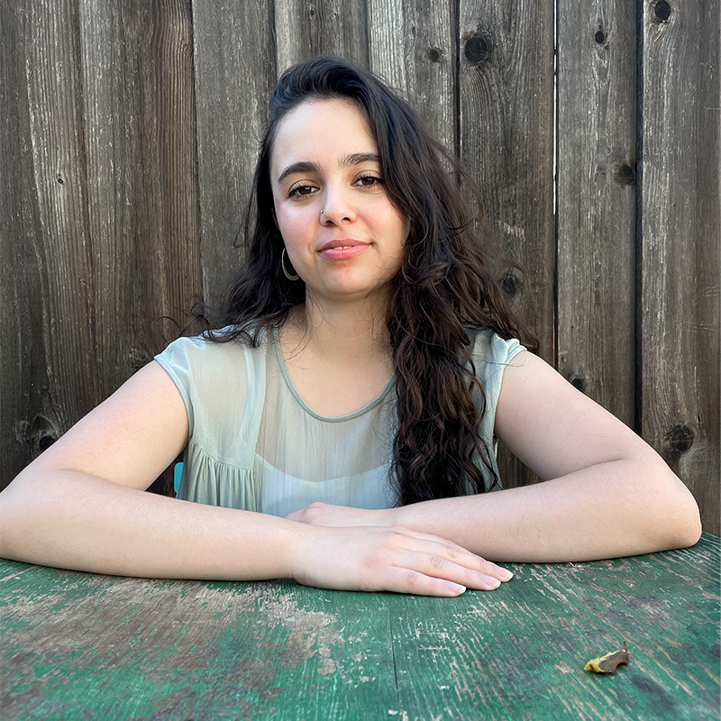 A woman with long dark hair sits at a green weathered table with her arms crossed, smiling slightly. She is wearing a light green, short-sleeved top, and is in front of a wooden fence.