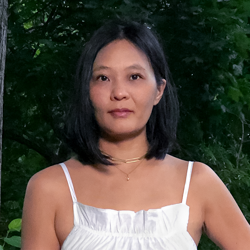 A woman with straight black hair wearing a white sleeveless top stands outdoors in front of green trees, looking directly at the camera with a neutral expression.