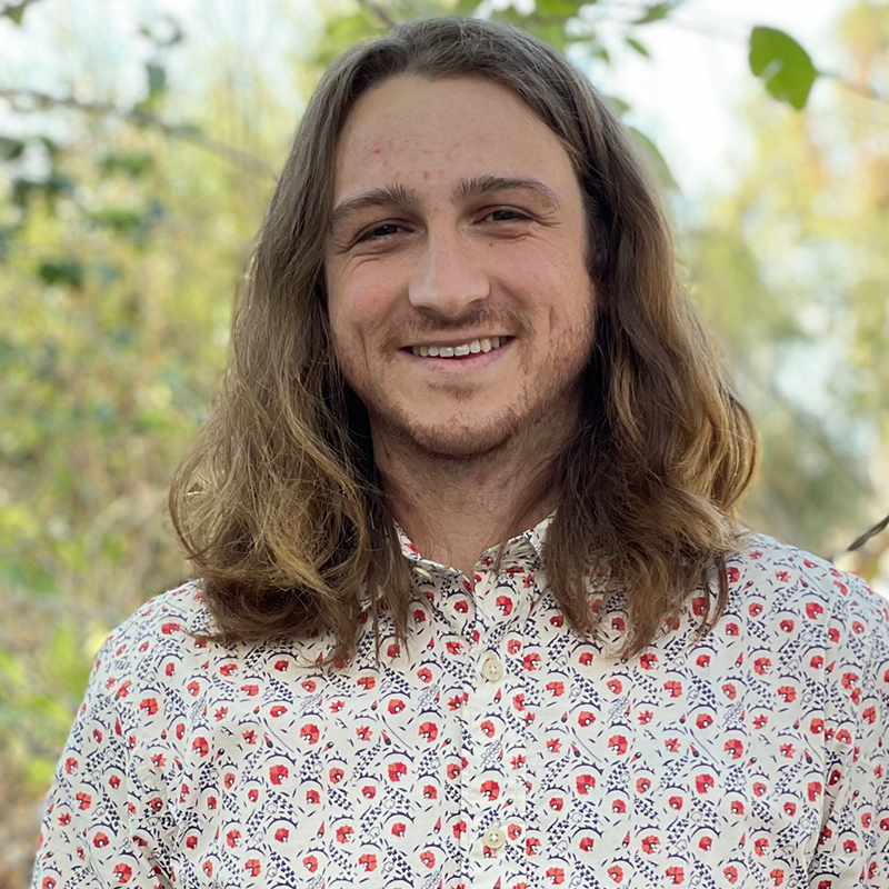 A person with long brown hair smiles outdoors, wearing a white shirt with a red and blue floral pattern. The background is filled with green foliage and soft sunlight.