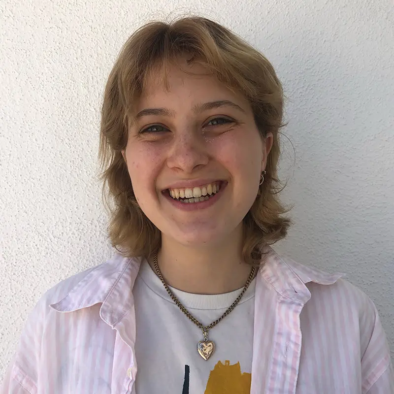 A person with short, light brown hair smiles widely, showing their teeth. They are wearing a white shirt, a light pink striped overshirt, and a heart-shaped pendant necklace, standing in front of a white textured wall.