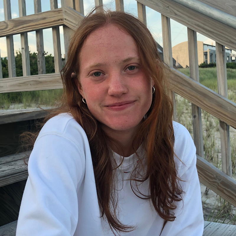 A young woman with long red hair and a white sweatshirt sits on wooden steps outdoors, smiling softly at the camera. Grassy dunes and buildings are visible in the background.