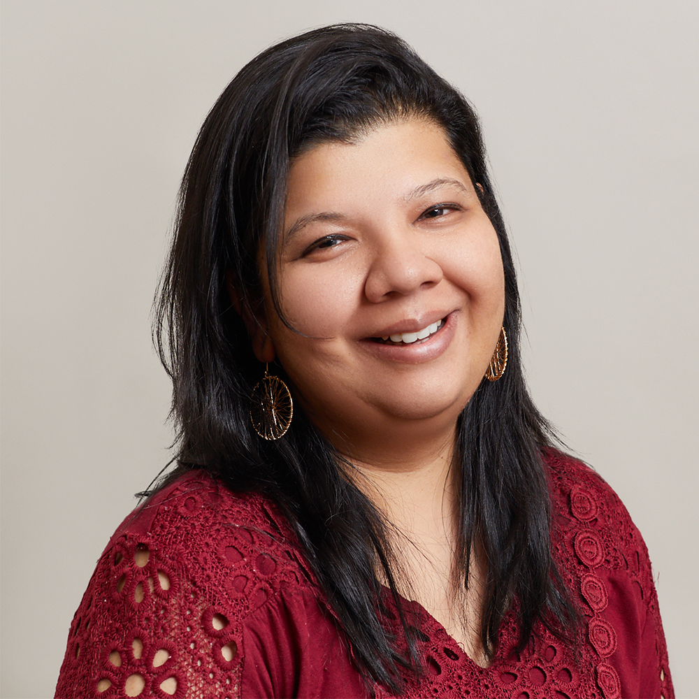 A woman with long dark hair, wearing gold hoop earrings and a burgundy top with lace details, smiles warmly at the camera against a plain light background.