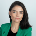 A woman with straight dark hair, wearing a green blazer over a black top and a delicate gold necklace, poses in front of a plain light background with a neutral expression, highlighting resilience in the face of social media online abuse.