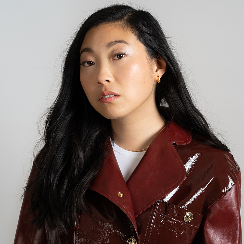 A woman with long black hair wearing a shiny maroon jacket over a white top looks at the camera with a neutral expression against a plain light background.