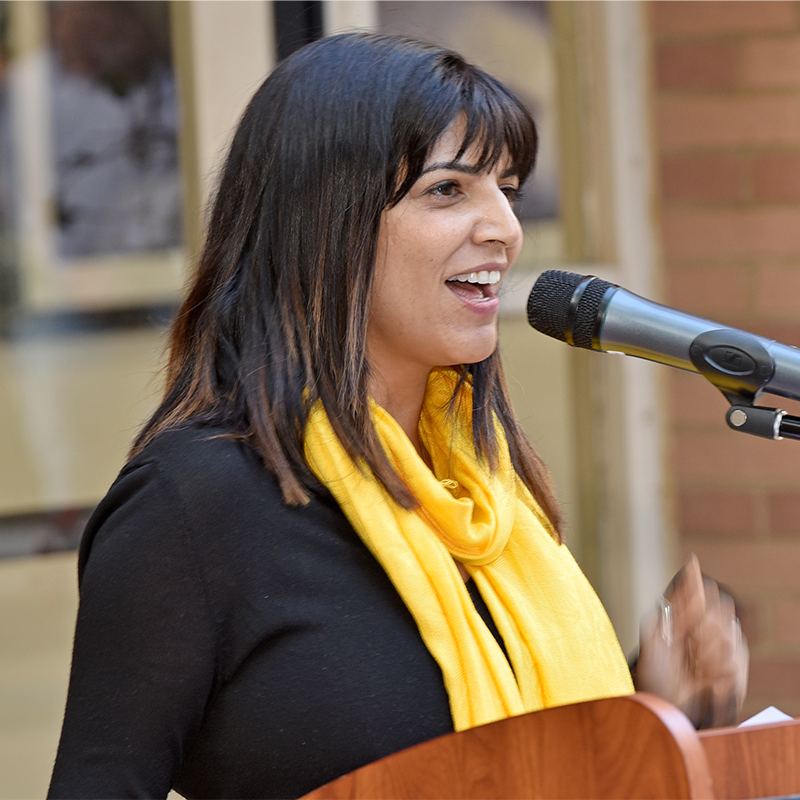 A woman with straight dark hair, wearing a yellow scarf and black top, speaks enthusiastically into a microphone at a podium.