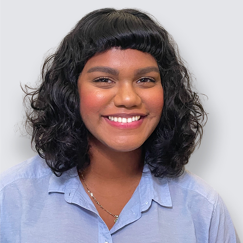 A woman with curly dark hair and bangs smiles at the camera. She wears a light blue collared shirt and a gold necklace, with a neutral gray background behind her.
