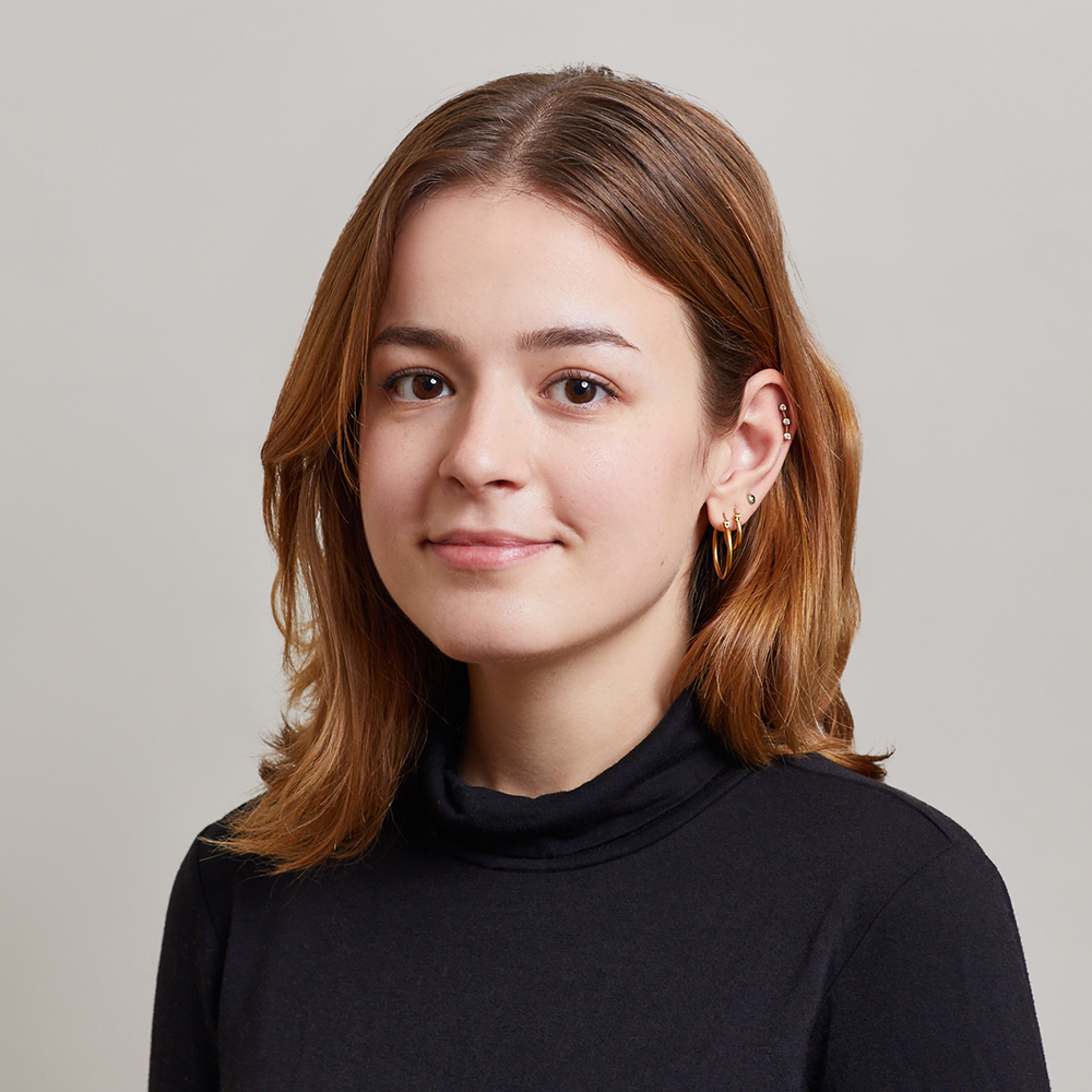 A young woman with shoulder-length brown hair wearing a black turtleneck and gold hoop earrings, smiling softly against a plain light grey background.