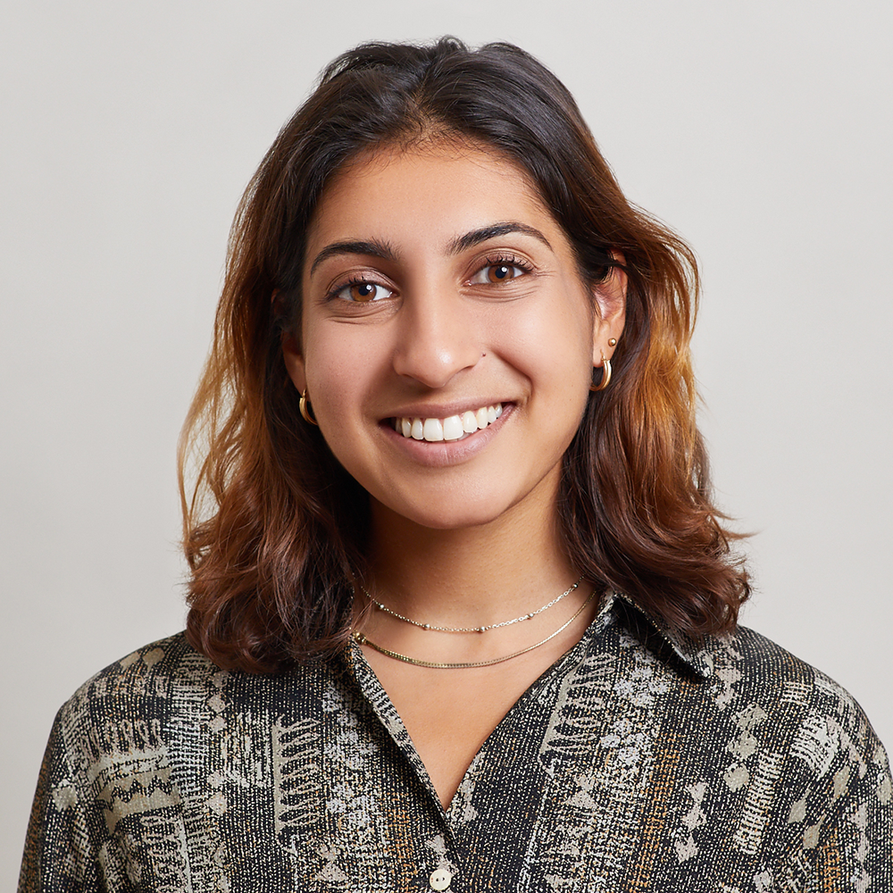 A young woman with shoulder-length brown hair smiles at the camera. She wears a patterned shirt, gold hoop earrings, and layered necklaces. The plain background highlights her confident style, reflecting the spirit of NYC Free Expression Advocacy Institute PEN.