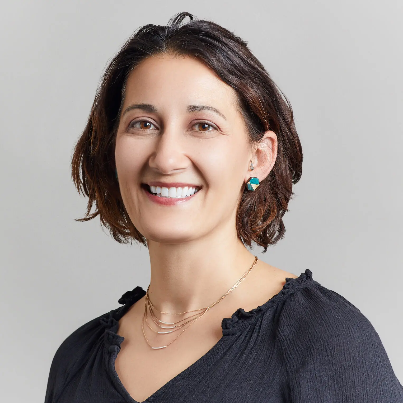 Smiling woman with short brown hair, wearing a black top, layered gold necklaces, and turquoise earrings, posed against a plain light gray background—capturing the spirit of NYC Free Expression Advocacy Institute PEN.