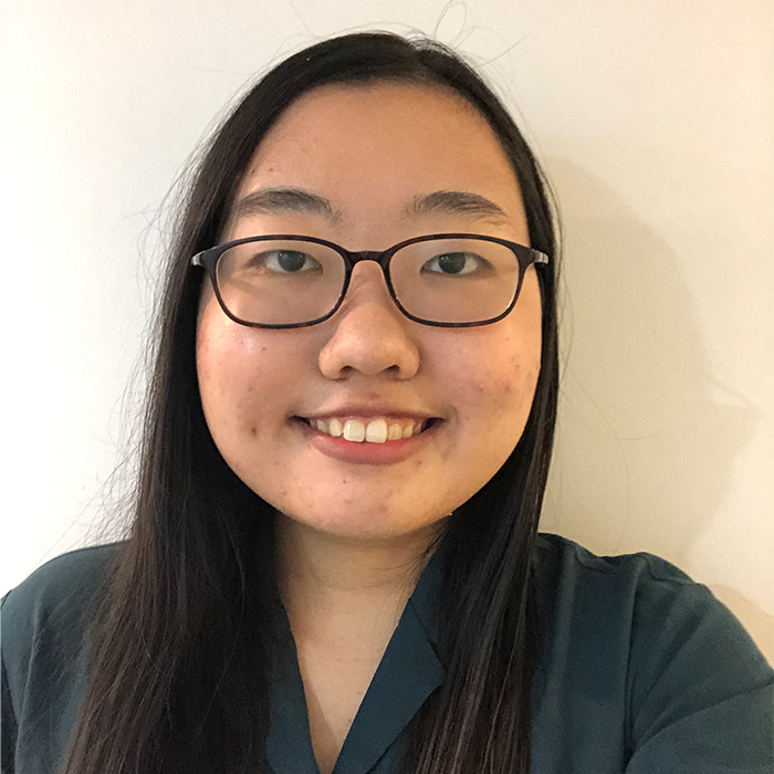 A young woman with long dark hair and glasses smiles at the camera. She is wearing a dark green top and stands against a plain white background.