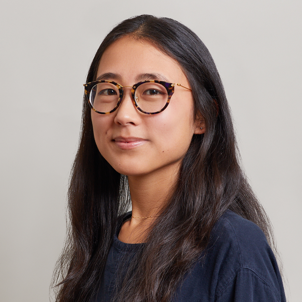 A young woman with long dark hair and glasses, wearing a black shirt, looks at the camera with a slight smile against a plain light background.