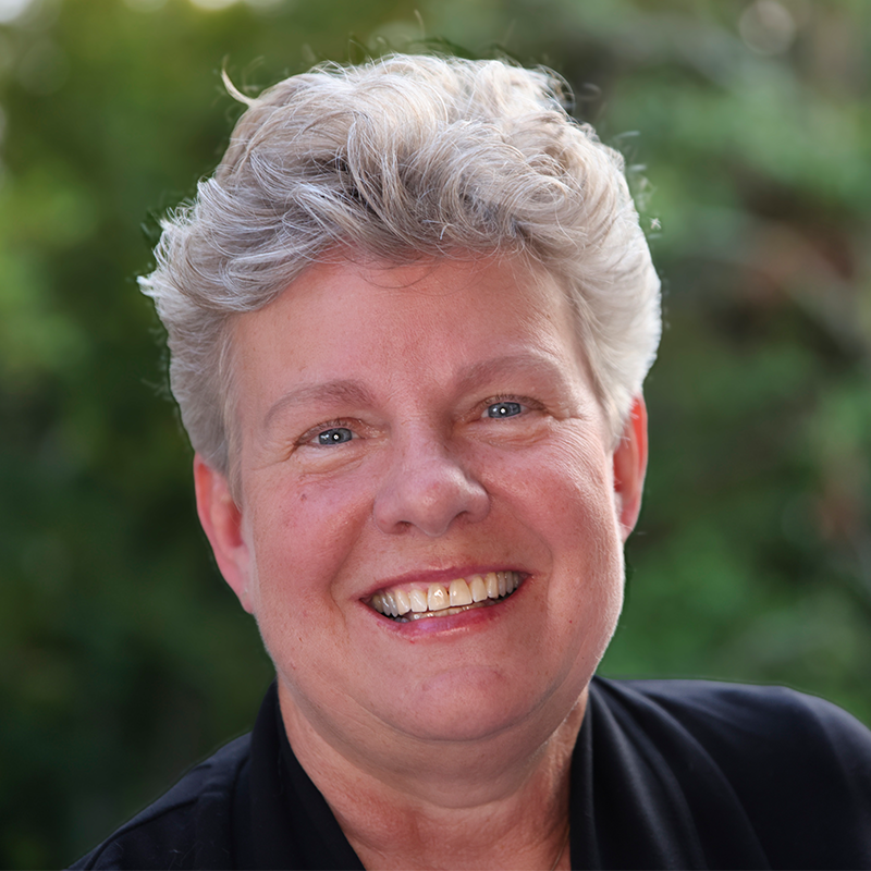 A smiling person with short, wavy gray hair wearing a dark top, posing outdoors with green foliage blurred in the background.