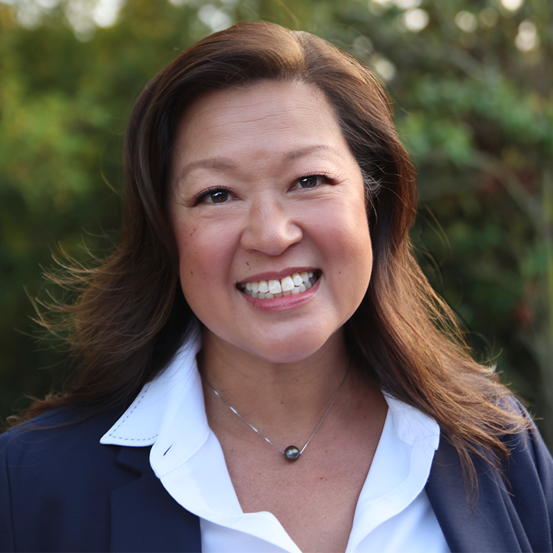 A woman with long brown hair, wearing a navy blazer over a white collared shirt and a silver necklace, smiles outdoors with trees and greenery in the background.